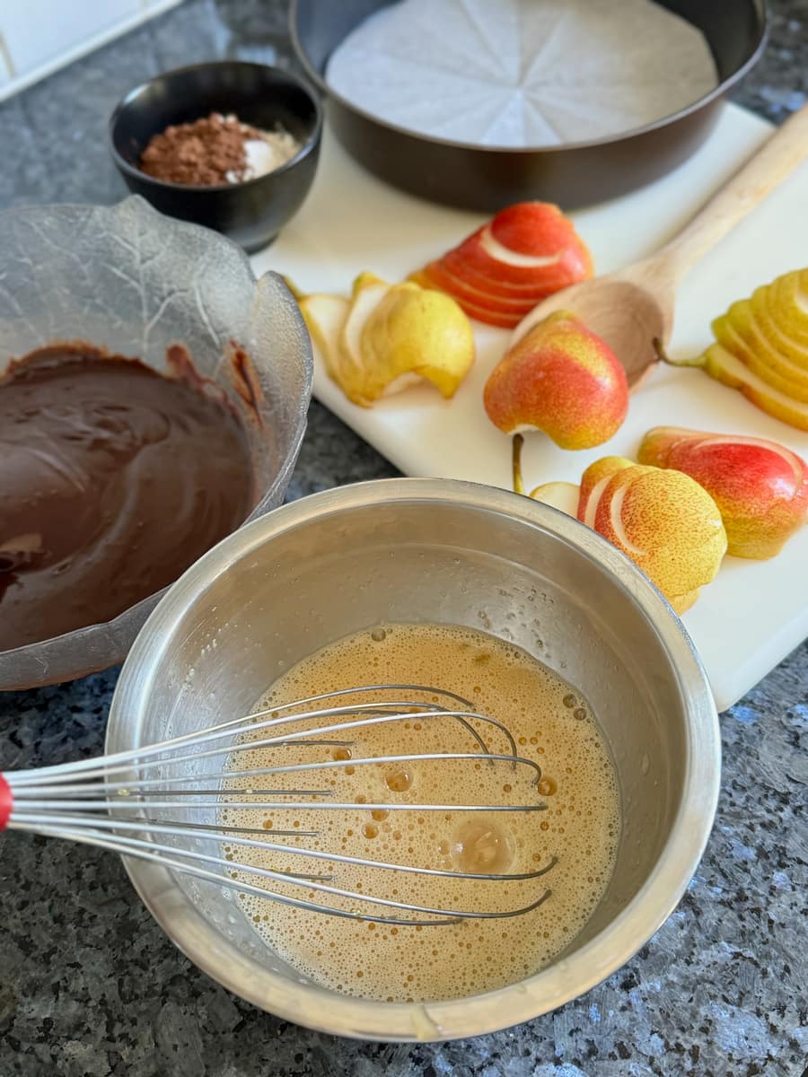 whisking eggs and sugar in a bowl next to another bowl of melted chocolate and butter with flour, a lined cake tin and sliced pears in the background